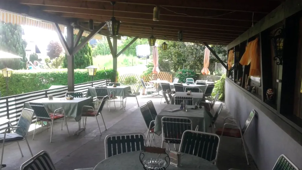 Überdachte Außenterrasse mit mehreren leeren Tischen und Stühlen, umgeben von üppigem Grün und Holzbalken.